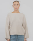 A woman with light hair tied back wears the Rag & Bone ingrid boat neck sweater—loose with chunky ribbing and wide sleeves—paired with blue jeans, standing against a plain light background.