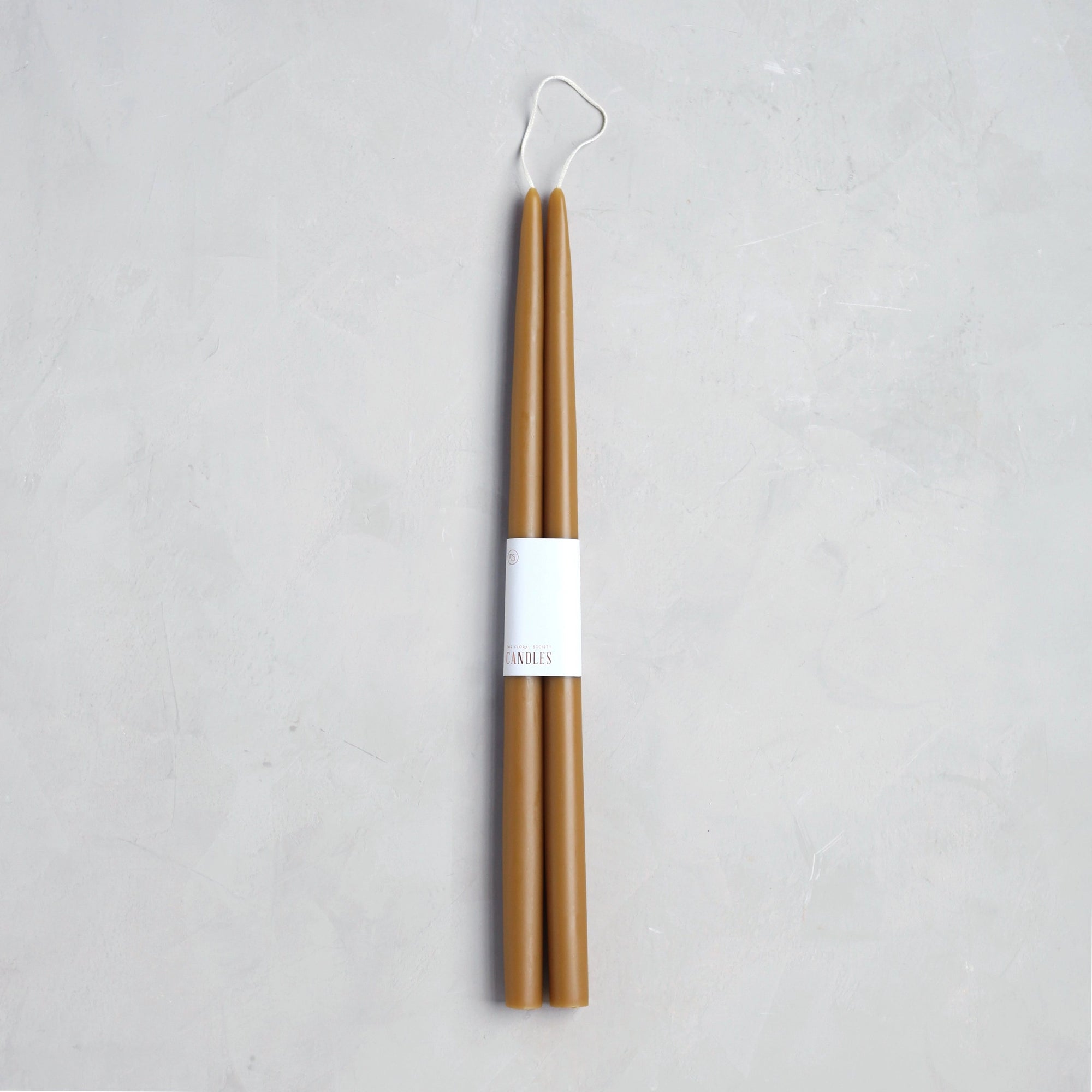 The Floral Society’s 18" Taper Candle - Pair is bundled with a white paper label and rests on a light gray textured surface—ideal for bringing elegantly styled décor to any space.