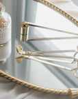 A glass bottle of apothecary matches and Faire’s Candle Wick Trimmer, along with two other gold accessories, sit on a braid-edged gold oval mirror atop a white cloth near a window.