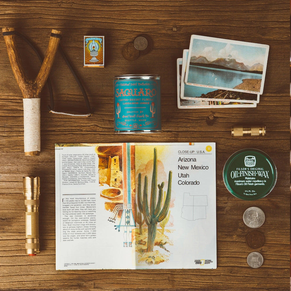A flat lay of vintage items on wood, featuring the Faire Saguaro National Park Candle, a slingshot, matches, blue can, postcards, flashlight, oil-finish wax, coins, and an open book with cactus florals and a Southwest US map.