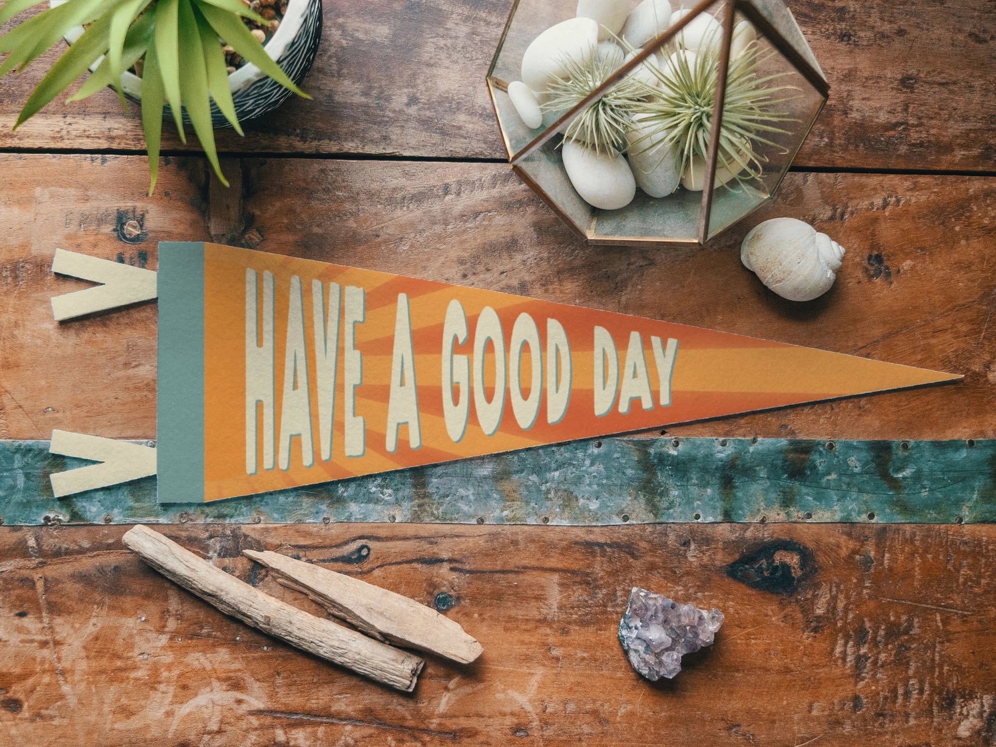 The Faire "Have a Good Day" Felt Pennant rests on a wooden surface amid plants, stones, and wood—this cheerful, motivational banner is perfect for brightening any space.