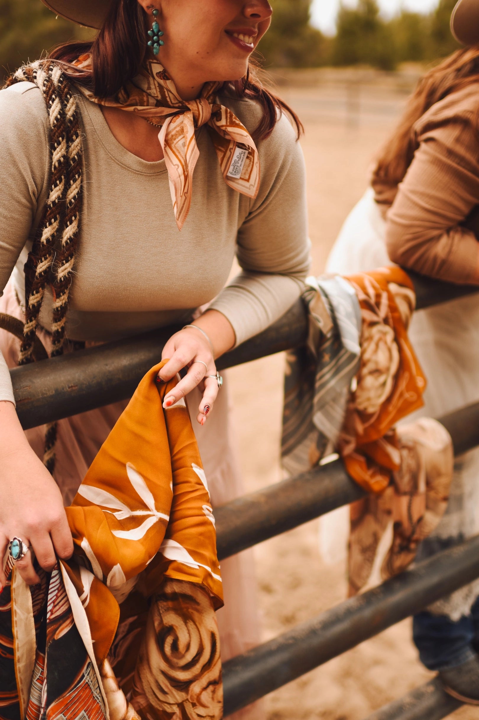 A woman in a tan long-sleeve shirt and Faire's Rustic Wild Rose Western Silk Scarf leans on a metal fence, holding warm-toned patterned fabrics—the perfect cowgirl accessory. Another person stands beside her outside, likely at a ranch or farm.
