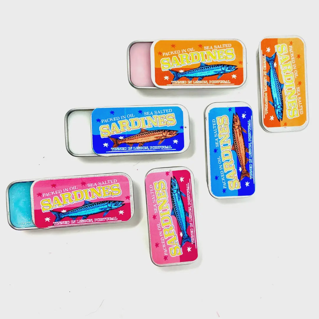 Faire's Sardine Tin Lip Balm is displayed on a white background, its vibrant, Portugal-inspired blue, orange, and pink packaging proudly showcasing "Sardine Tin Lip Balm." Some tins are open, revealing the enticing charm of their contents.