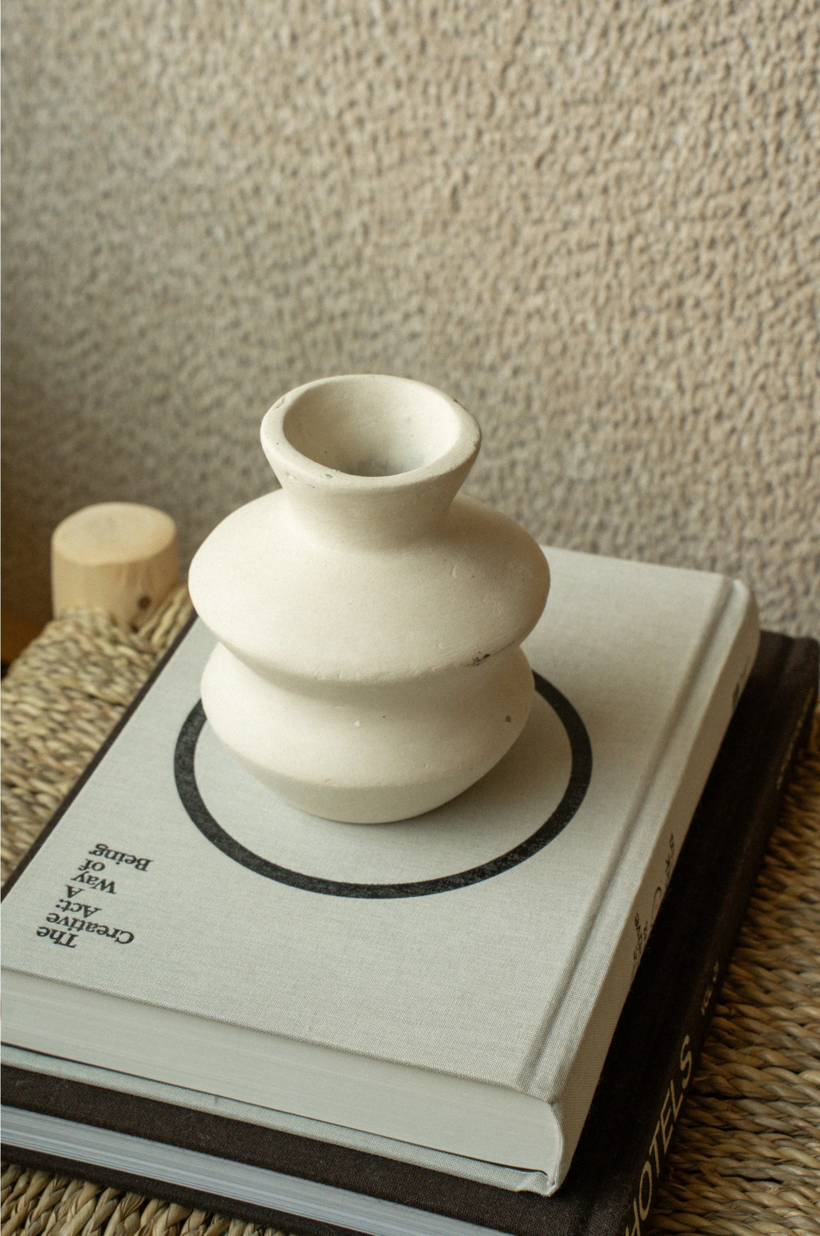 The Indaba Wave Paper Mache Bud Vase, handcrafted and petite, sits atop a minimalist-covered book on a woven mat against a textured beige wall—ideal for sustainable home decor.