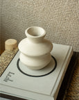 The Indaba Wave Paper Mache Bud Vase, handcrafted and petite, sits atop a minimalist-covered book on a woven mat against a textured beige wall—ideal for sustainable home decor.