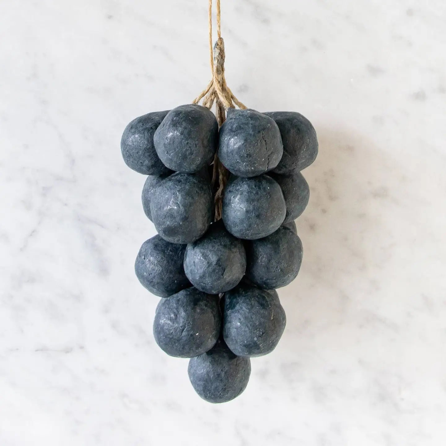 Handcrafted Grapes Soap by Faire, resembling a cluster of dark grapes, hangs by twine against a white marble background for an artisanal touch.