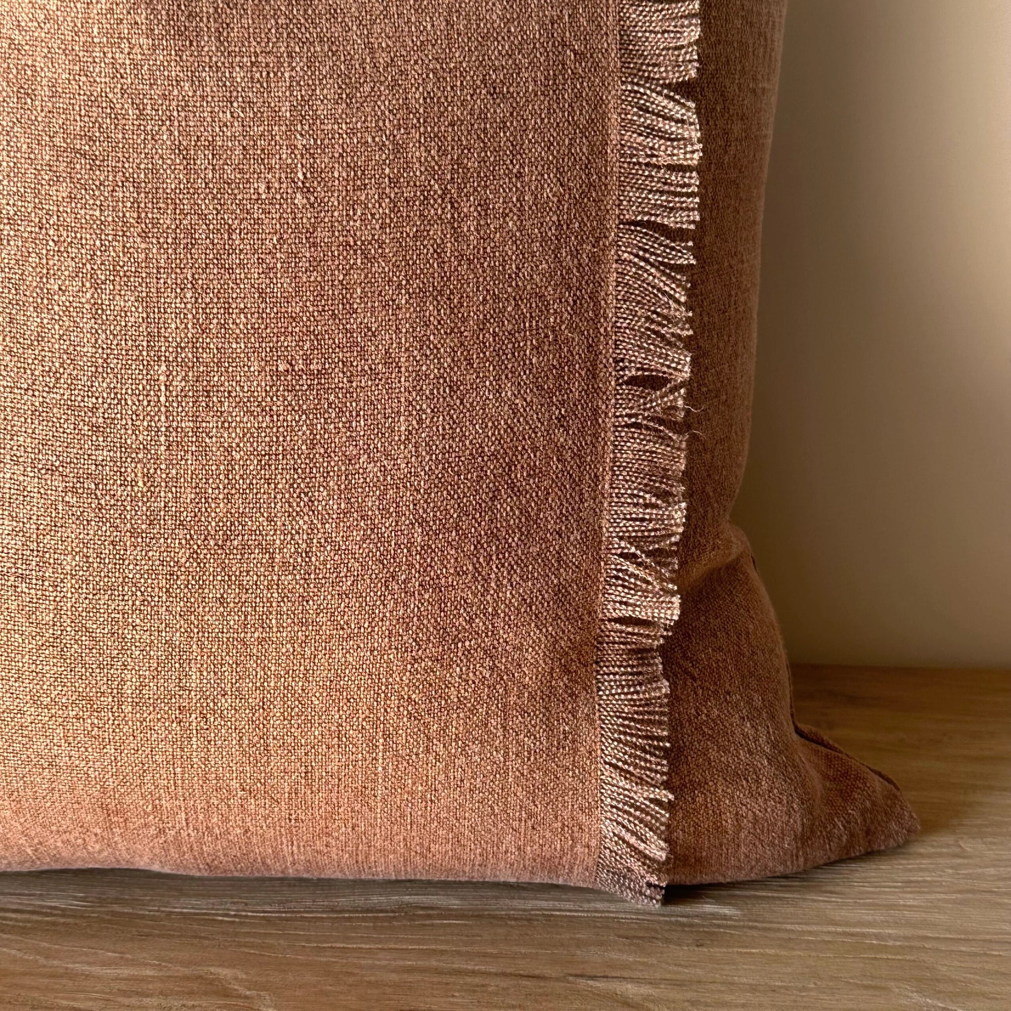 The Fringe Accent Pillow Havane 22x22 features a rust brown hue with fringe detailing along the right edge, shown close-up as it rests on a wooden surface against a light beige wall.