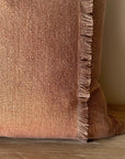 The Fringe Accent Pillow Havane 22x22 features a rust brown hue with fringe detailing along the right edge, shown close-up as it rests on a wooden surface against a light beige wall.