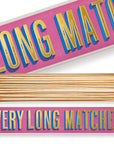 A rectangular matchbox features "Long Fireplace Matches" by Faire in bold, colorful letters on a pink background. The open box reveals neatly arranged long wooden matches with Magnifique white tips.