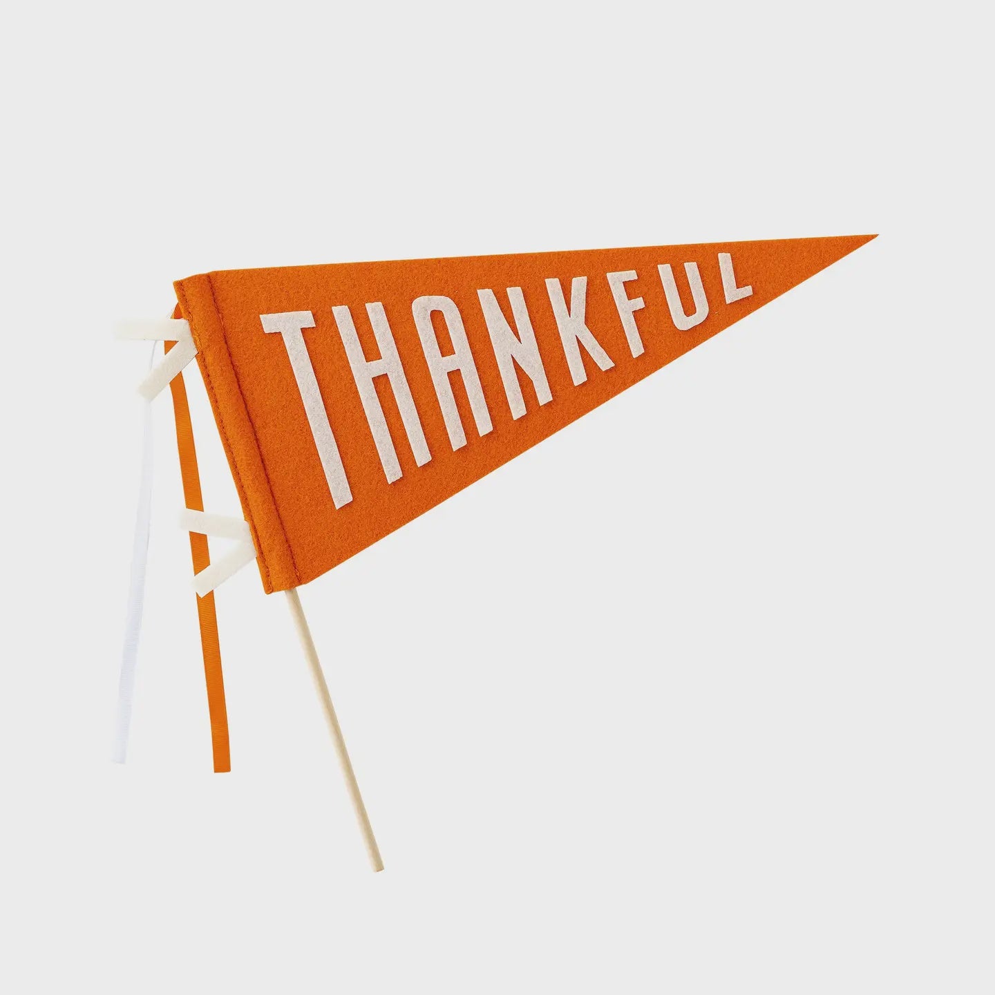 The Harvest Thankful Felt Pennant by Faire is adorned with large white letters spelling "THANKFUL" across an orange background. Affixed to wooden sticks, it serves as a perfect festive centerpiece against a plain white backdrop, embodying the spirit of Thanksgiving.