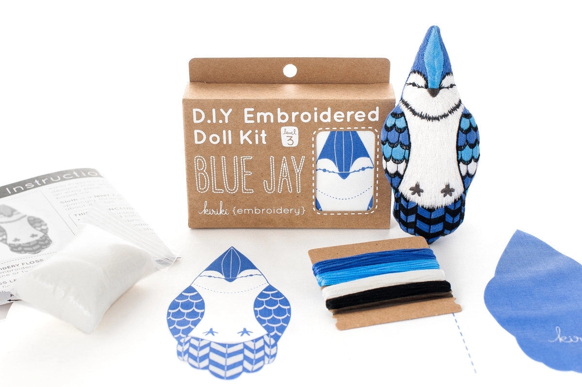 The Faire Blue Jay Embroidery Kit includes a box, pre-printed fabric, colored threads (blue, black, white), stuffing, sewing instructions, and lets you make a charming embroidered Blue Jay doll.
