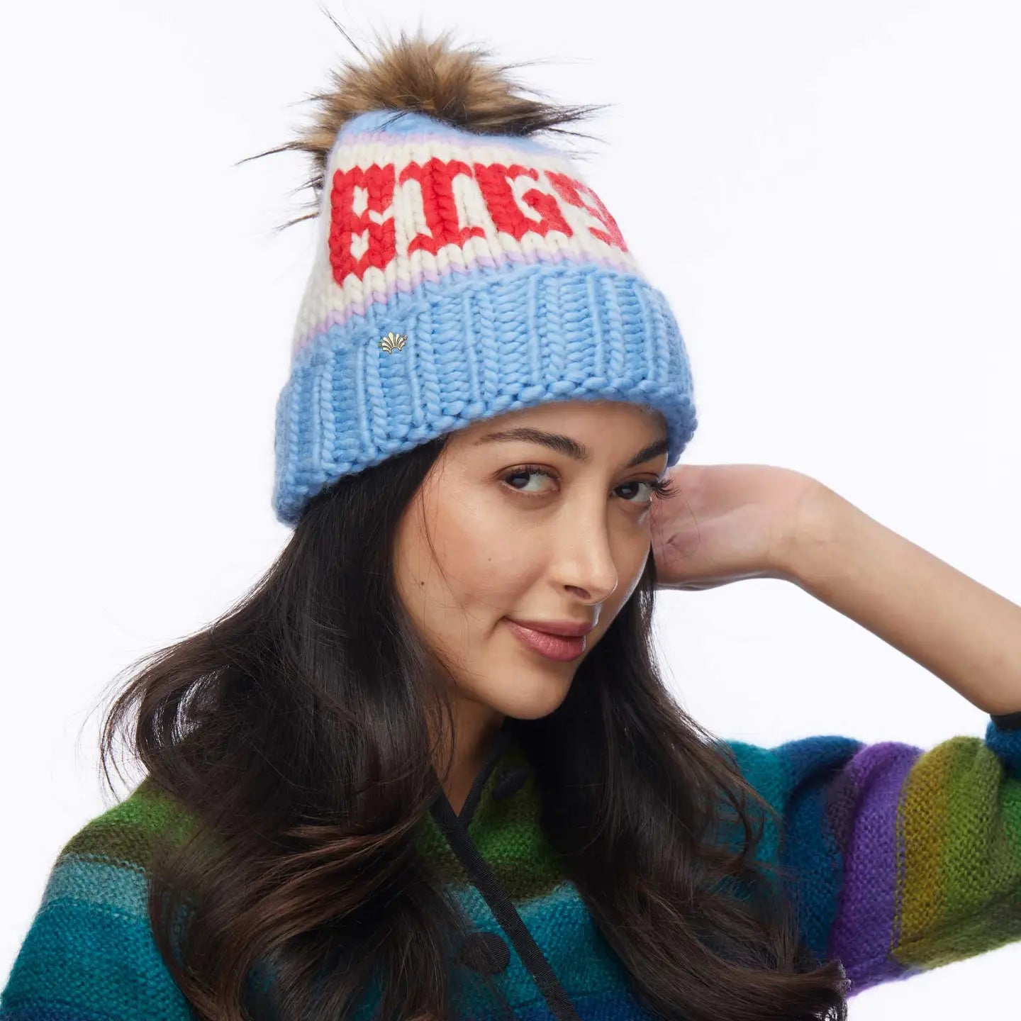A woman with long dark hair wears the Faire Knit Ski Hat, a luxury merino wool beanie with a faux fur pom pom, paired with a striped multicolored sweater, posing against a plain background.