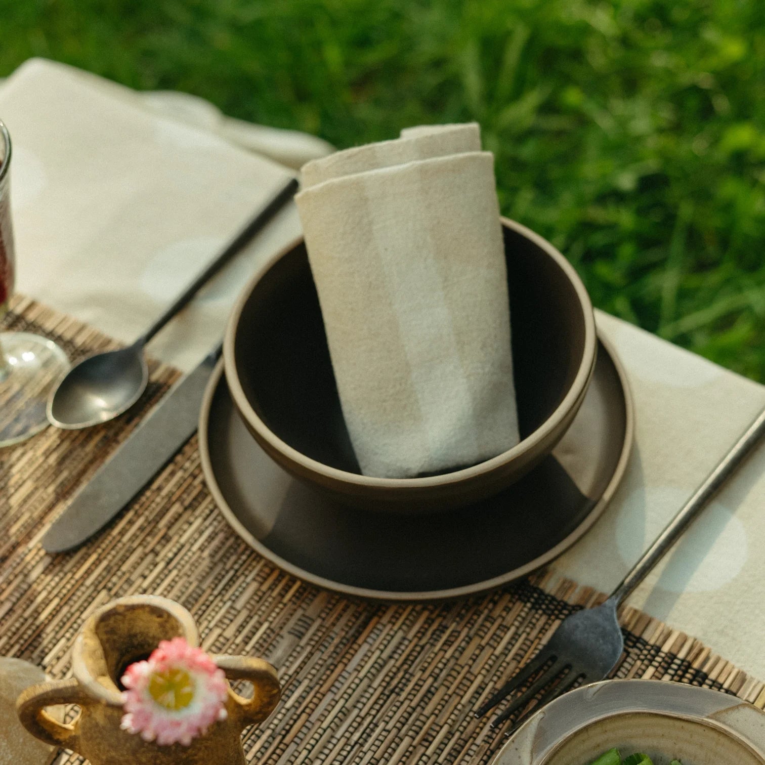An Indaba 4-7122 Stripe Resist Napkin is neatly folded in a dark bowl on a matching plate, with fork, spoon, and knife set on a woven placemat atop an outdoor table against a grassy backdrop.
