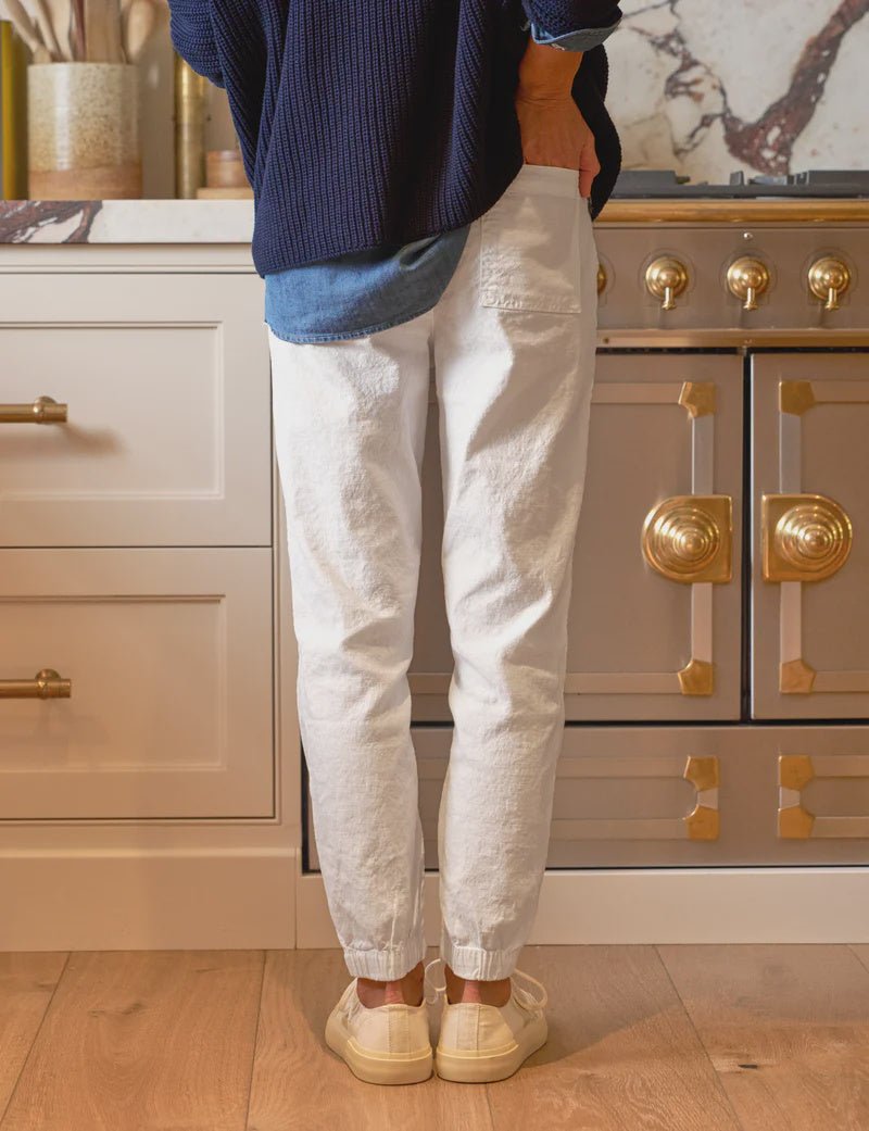 In a kitchen with a stove featuring ornate gold accents, someone in a blue sweater, denim shirt, and white pants paired with beige sneakers radiates casual elegance. Complemented by warm wood floors and cabinets, their look is enhanced by the practicality of the Frank & Eileen Jameson Utility Jogger.