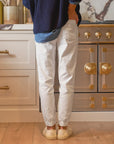 In a kitchen with a stove featuring ornate gold accents, someone in a blue sweater, denim shirt, and white pants paired with beige sneakers radiates casual elegance. Complemented by warm wood floors and cabinets, their look is enhanced by the practicality of the Frank & Eileen Jameson Utility Jogger.