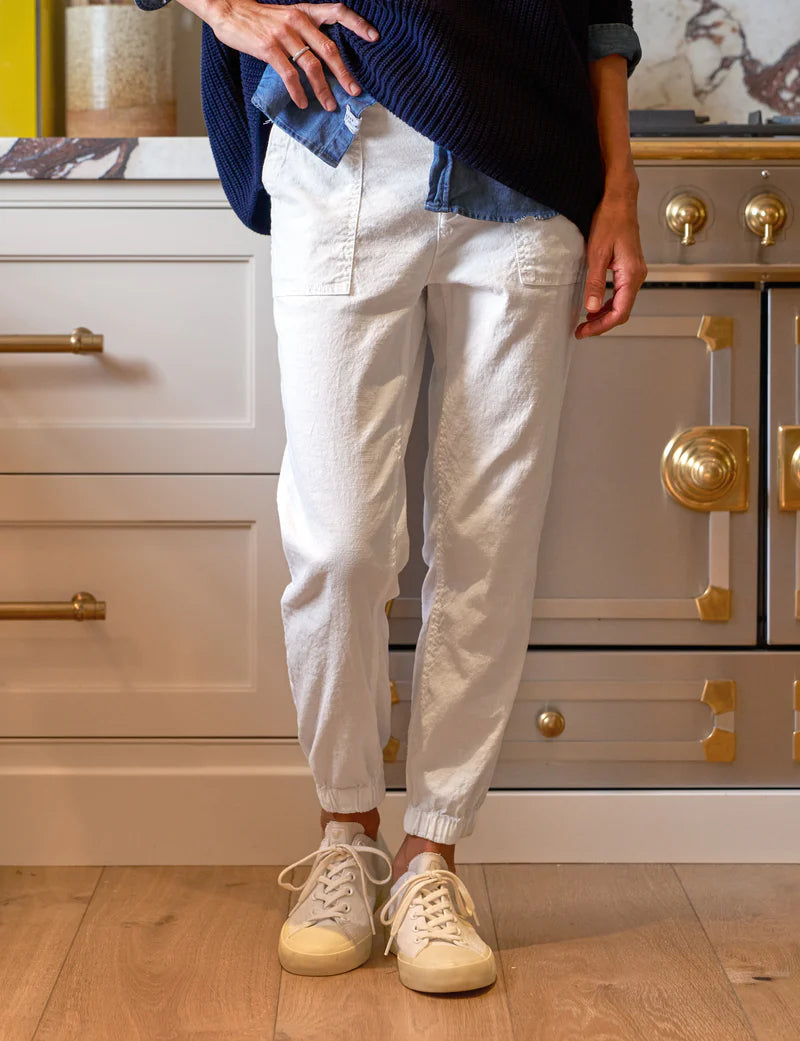 Someone wearing Frank & Eileen's Jameson Utility Jogger in white and cream sneakers is standing in a kitchen with white cabinets and a stove with gold accents. Their upper body shows they are dressed in a dark blue sweater over a denim shirt.