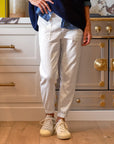 Someone wearing Frank & Eileen's Jameson Utility Jogger in white and cream sneakers is standing in a kitchen with white cabinets and a stove with gold accents. Their upper body shows they are dressed in a dark blue sweater over a denim shirt.