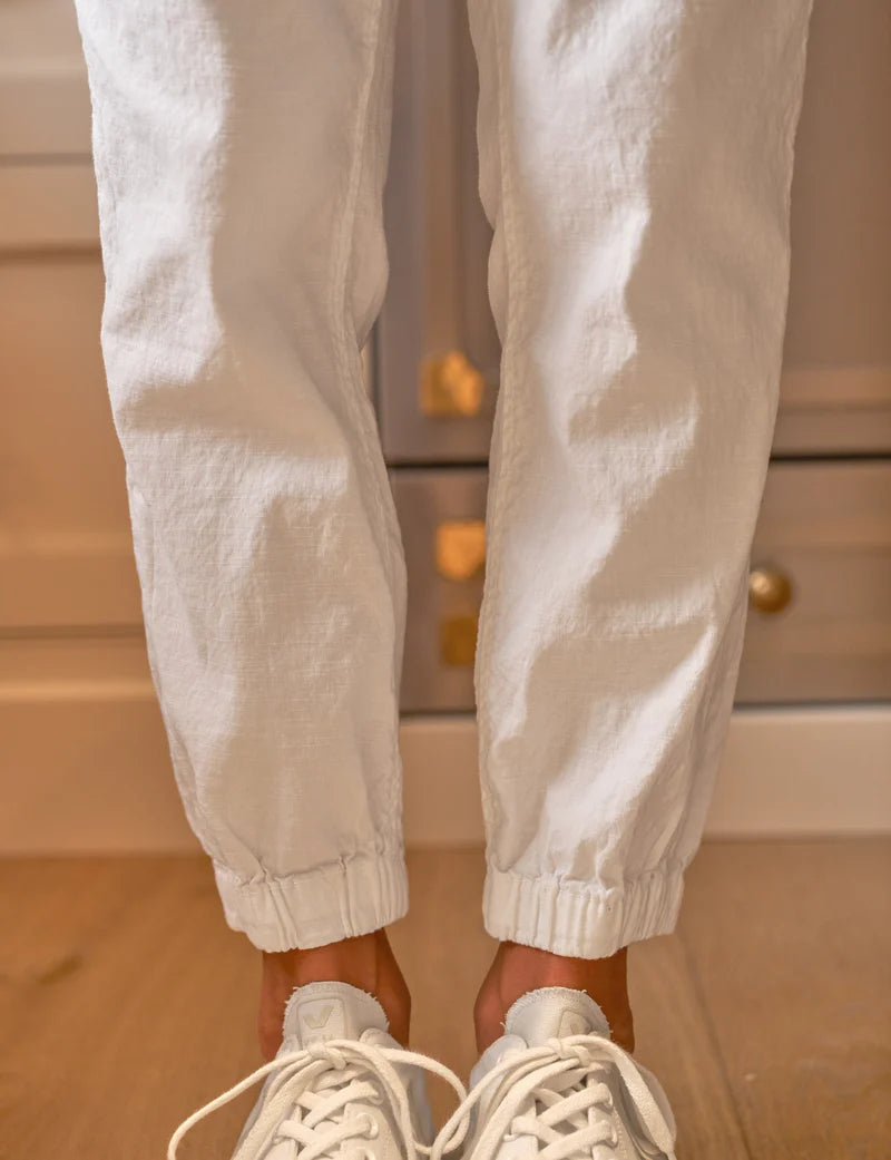 A person stands on a wooden floor wearing white sneakers and Frank & Eileen's Jameson Utility Jogger with utility pockets. The background features a light-colored, textured cabinet with brass knobs.