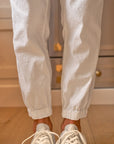 A person stands on a wooden floor wearing white sneakers and Frank & Eileen's Jameson Utility Jogger with utility pockets. The background features a light-colored, textured cabinet with brass knobs.