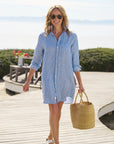 A woman in the Frank & Eileen MARY Shirtdress S25, styled with sunglasses, sandals, and a woven straw bag, strolls a beachside boardwalk framed by flowers and greenery.