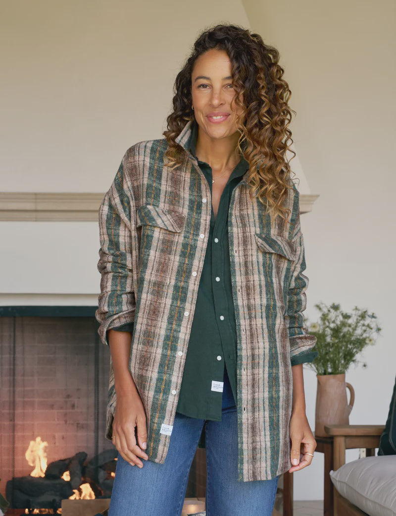 A woman with curly hair smiles indoors, wearing Frank & Eileen’s McLoghlin Utility Shirt under a brown and green plaid flannel and blue jeans. A lit fireplace and a small table with a plant are in the background, capturing relaxed California-cool style.