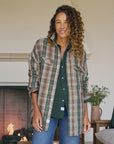A woman with curly hair smiles indoors, wearing Frank & Eileen’s McLoghlin Utility Shirt under a brown and green plaid flannel and blue jeans. A lit fireplace and a small table with a plant are in the background, capturing relaxed California-cool style.