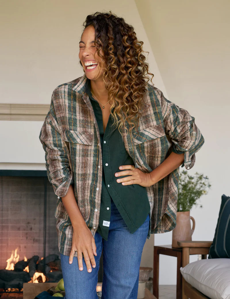 A woman with curly hair, dressed in a green shirt and the Frank & Eileen McLoghlin Utility Shirt, smiles and laughs indoors by a lit fireplace. A vase and cushions sit on the side table behind her.