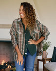 A woman with curly hair, dressed in a green shirt and the Frank & Eileen McLoghlin Utility Shirt, smiles and laughs indoors by a lit fireplace. A vase and cushions sit on the side table behind her.