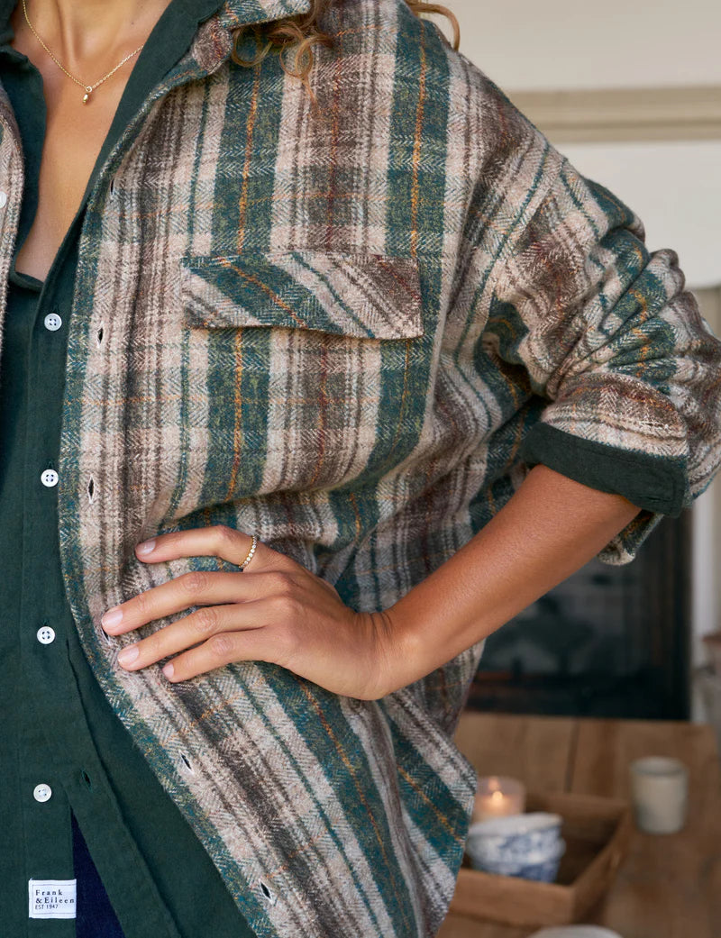 Wearing the Frank & Eileen McLoghlin Utility Shirt—an oversized green plaid button-up—over a dark green top, a person stands with one hand on their hip. The focus is on California-cool layers against a cozy indoor backdrop.