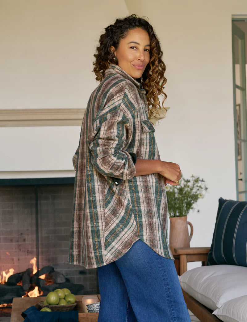 A woman with curly hair wears the Frank & Eileen McLoghlin Utility Shirt and blue jeans, smiling indoors near a lit fireplace. A bowl of apples and flowers sit on a table by a sofa with blue pillows.