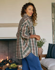 A woman with curly hair wears the Frank & Eileen McLoghlin Utility Shirt and blue jeans, smiling indoors near a lit fireplace. A bowl of apples and flowers sit on a table by a sofa with blue pillows.