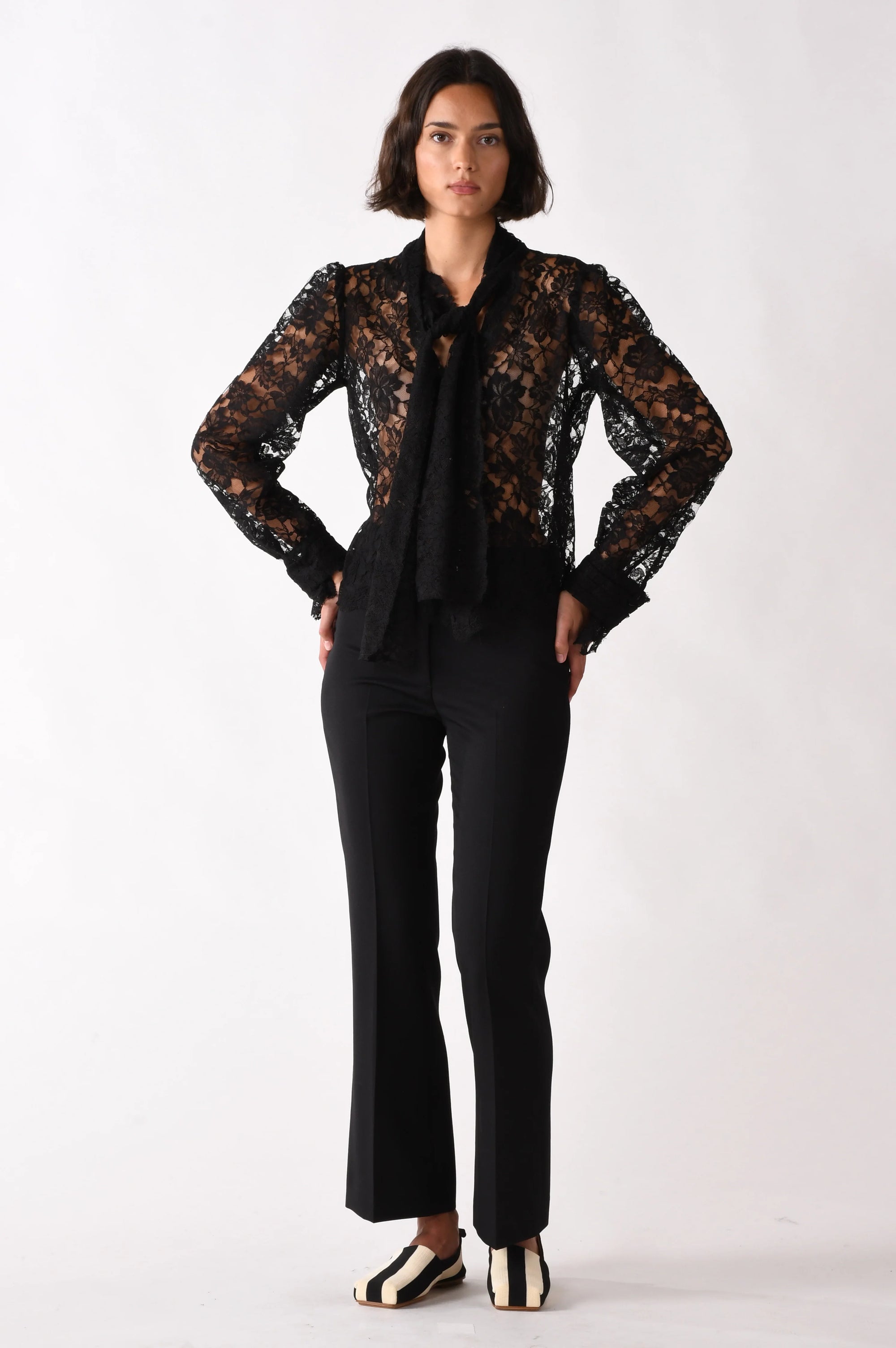 A person stands against a white background wearing Cissa's The Bow Tie Shirt—a black lace blouse with a scalloped hem—paired with black pants and white shoes with black accents. Hands on hips, they have a neutral expression.