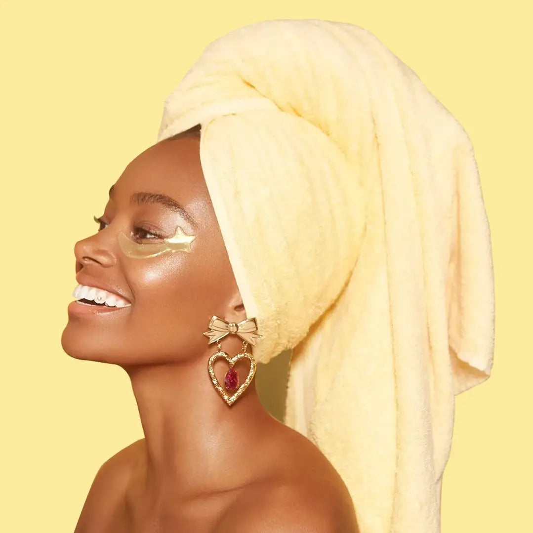 A woman with glowing skin beams, showcasing the magic of Faire's Collagen Star Eye Mask. A yellow towel crowns her head, and under-eye patches complement her heart-shaped earrings with red gems. Against a matching yellow background, she radiates bright, harmonious elegance.