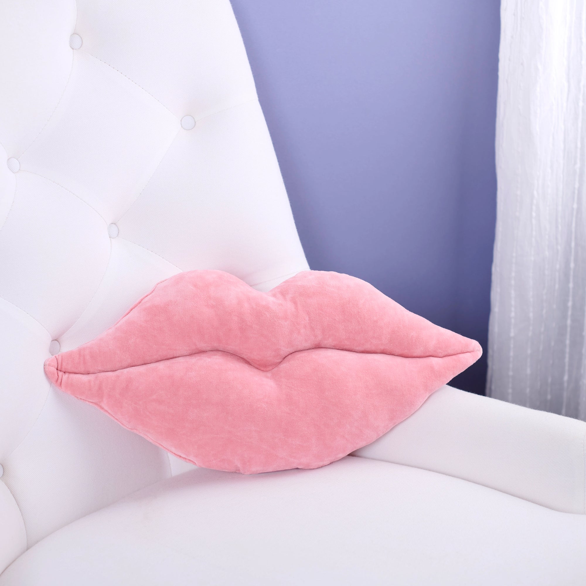 The Faire Lip Pillow, shaped like pink lips, sits on a white tufted chair with a lavender wall and sheer curtain in the background—a playful accent to add charm to any space.