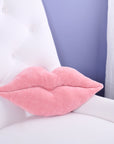 The Faire Lip Pillow, shaped like pink lips, sits on a white tufted chair with a lavender wall and sheer curtain in the background—a playful accent to add charm to any space.
