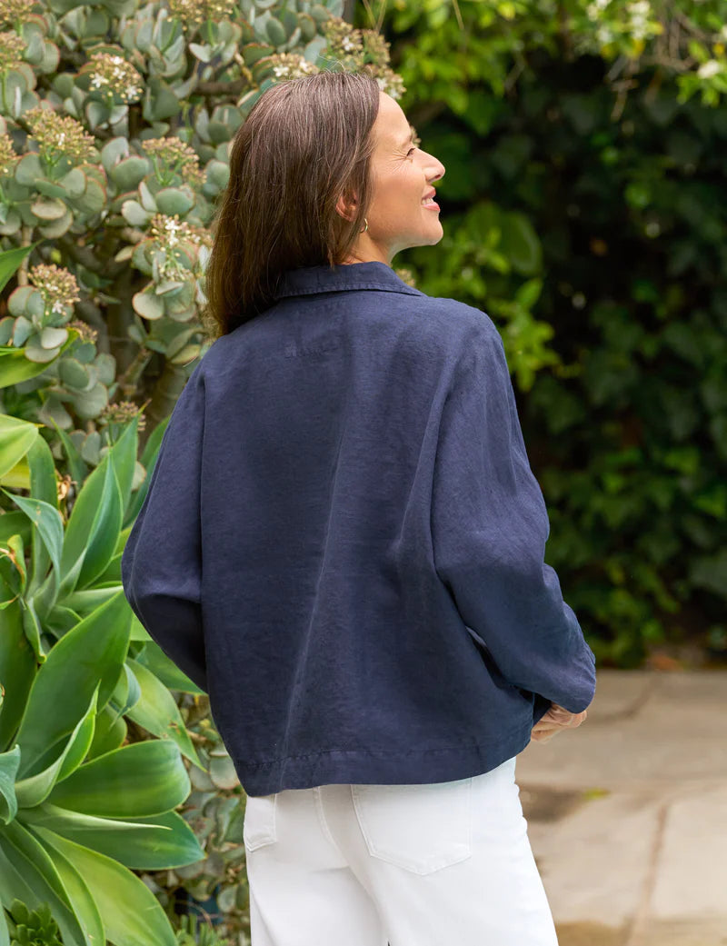 A woman with long brown hair stands outdoors, smiling amid lush greenery, wearing the Frank & Eileen Gabi Popover Shirt and white pants, exuding laidback elegance with her back to the camera.