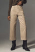 A person wears the Denimist Georgia Work Pant—beige, high-waisted, wide-leg pants with front patch pockets—with a brown long-sleeve top and black ankle boots, standing against a plain gray background.