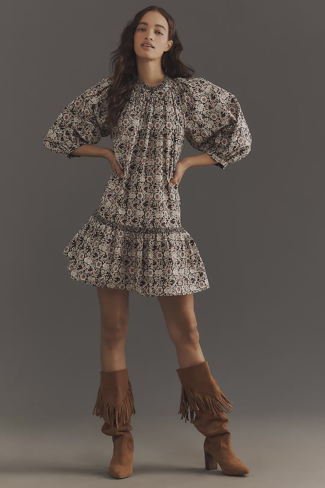 A person confidently stands with hands on hips wearing a Rhode Angelica mini dress featuring puffed sleeves. Made from organic cotton with a floral pattern, it pairs perfectly with fringed brown suede boots. The neutral gray background enhances their poise.