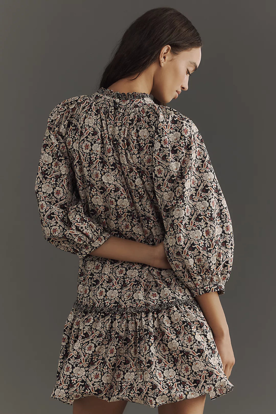 A woman with long dark hair poses against a plain gray background, looking to her right. She wears Rhode's Angelica Dress, a floral mini dress with puffed sleeves, a smocked waist, and a braided neckline made from organic cotton. One hand rests on her back.