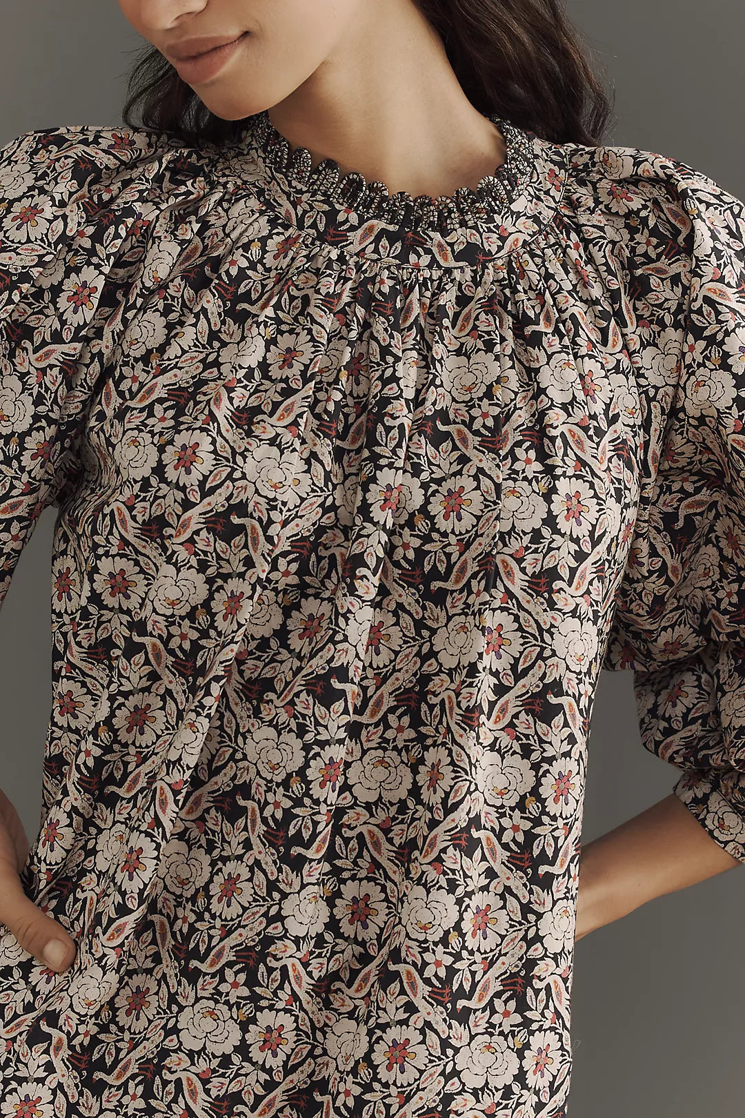 Wearing an Angelica Dress by Rhode in a floral pattern with puffed sleeves, a woman showcases standout style. The organic cotton dress has a gathered neckline in beige, red, and dark tones. She rests her hand on her hip against a plain gray background.