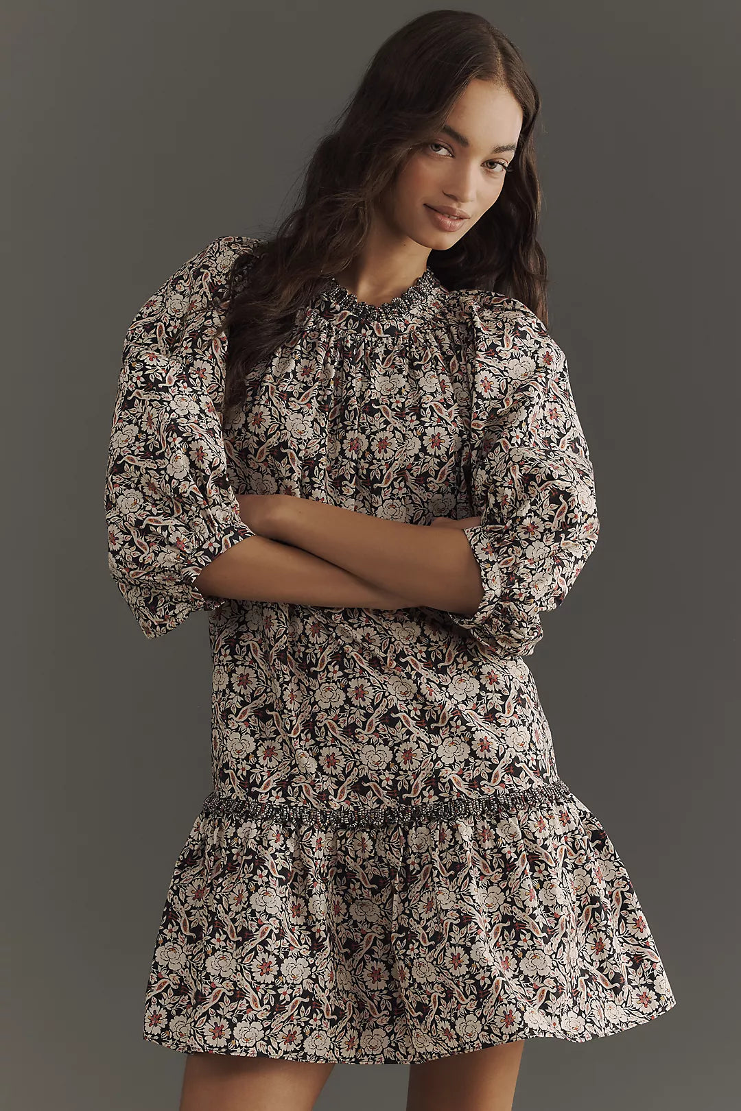 The woman models Rhode's Angelica Dress, a mini style featuring a patterned, long-sleeve design with a ruffled hem, made from soft organic cotton. She stands against a gray background, her long wavy hair framing her face as she gives a slight smile.