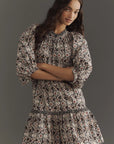 The woman models Rhode's Angelica Dress, a mini style featuring a patterned, long-sleeve design with a ruffled hem, made from soft organic cotton. She stands against a gray background, her long wavy hair framing her face as she gives a slight smile.