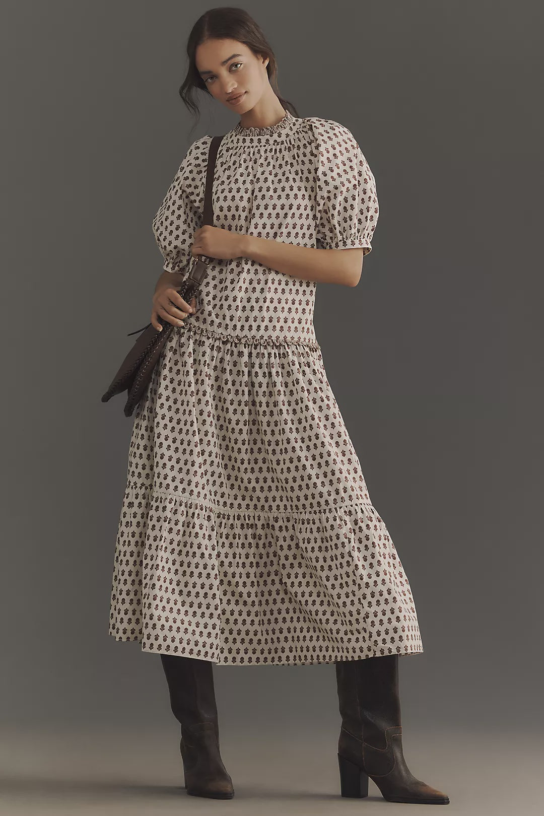 A person poses against a plain backdrop wearing the Rhode Aderyn Dress, featuring a floral print, puffed sleeves, and a tiered skirt. They complete the look with dark knee-high boots and a handbag. Standing with one foot forward adds to the outfit's casual elegance.