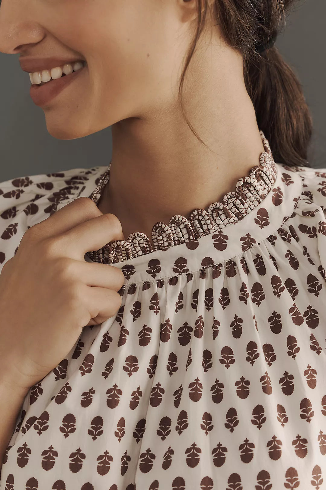 A woman smiles in a beige Aderyn Dress by Rhode, featuring a brown floral print and intricate beadwork on the collar. The background is a solid neutral tone.