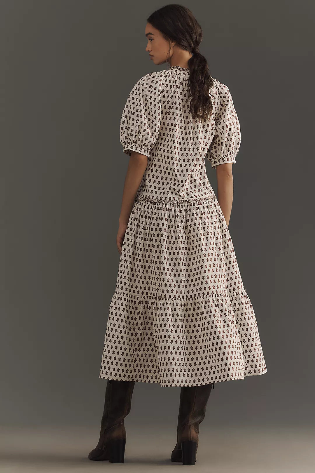 A woman stands sideways in the Aderyn Dress by Rhode, featuring a long floral print with puff sleeves and a ruffle hem. Her hair is tied in a loose ponytail, and she wears knee-high boots against a neutral backdrop.