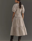 A woman stands sideways in the Aderyn Dress by Rhode, featuring a long floral print with puff sleeves and a ruffle hem. Her hair is tied in a loose ponytail, and she wears knee-high boots against a neutral backdrop.