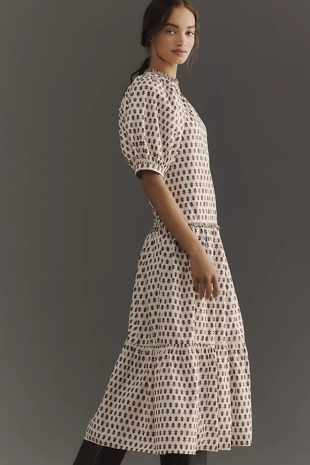 A woman in a Rhode Aderyn Dress stands sideways against a gray background. The patterned, ankle-length dress features puffed sleeves and a ruffle hem. Her dark hair is tied back, and the floral print enhances her calm expression.