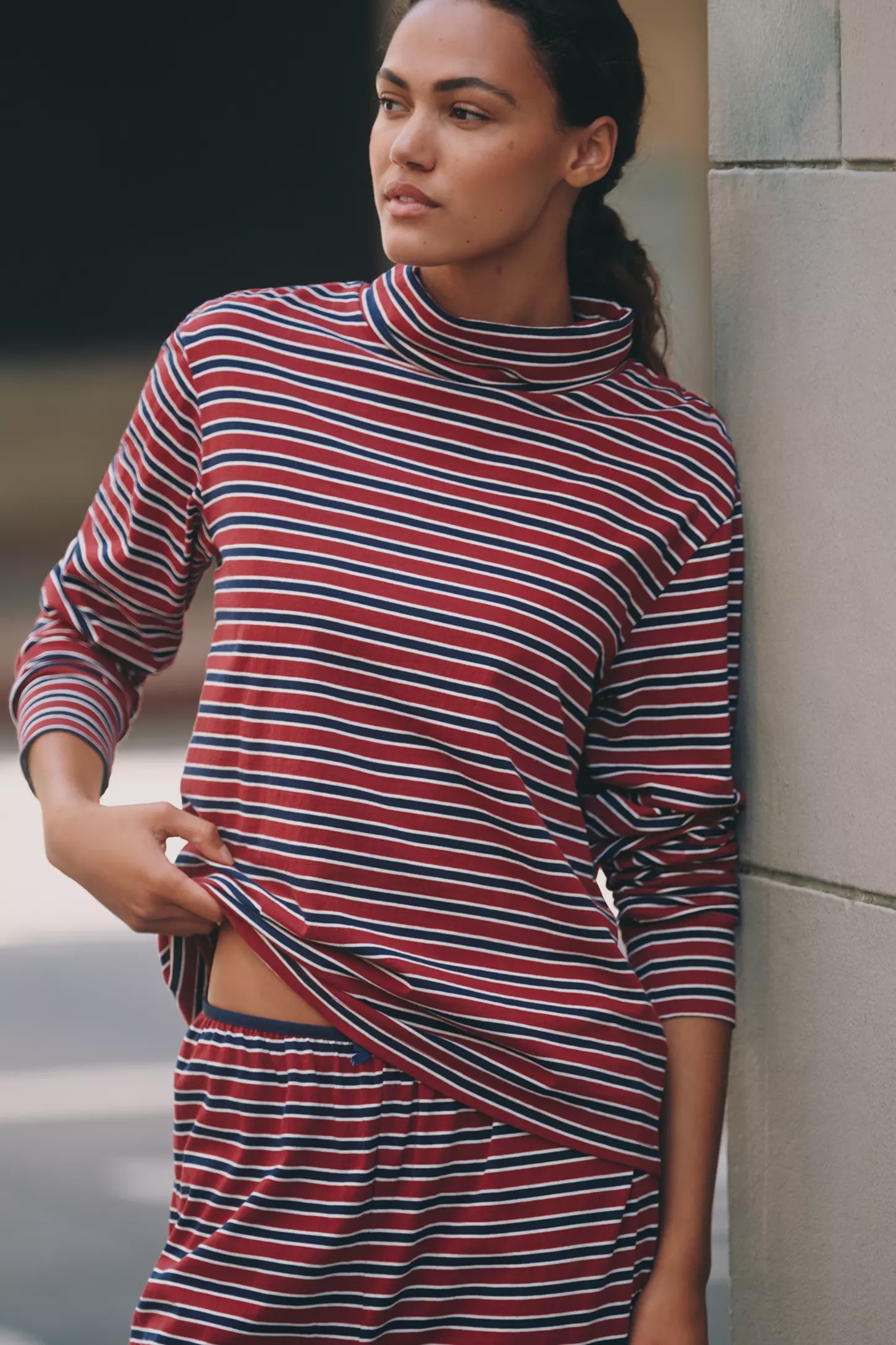 A woman in Donni’s Striped Jersey Turtleneck, featuring red, white, and blue horizontal stripes, stands against a light wall and looks to the side.