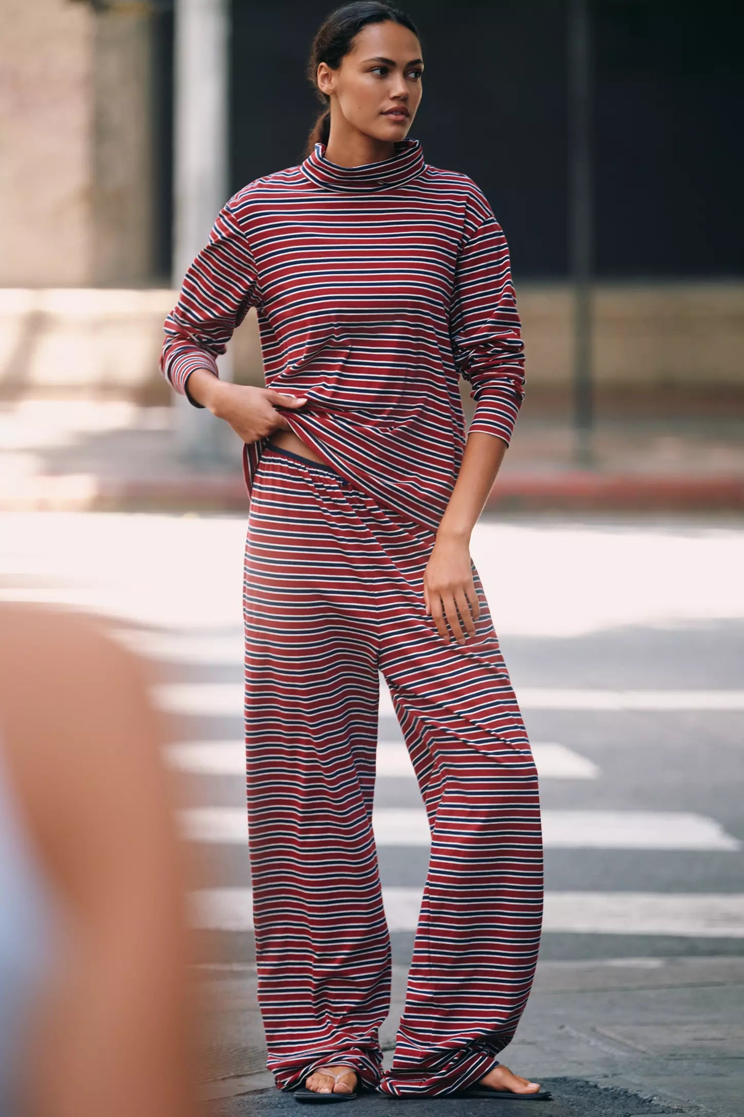A person stands on a city street wearing Donni's The Striped Jersey Turtleneck with matching wide-leg pants and sandals, gazing calmly to the side.