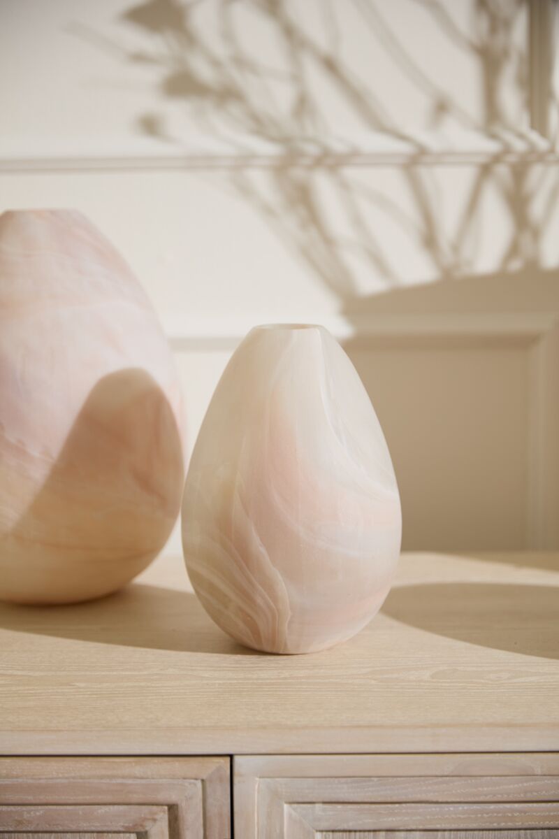 A smooth, pale pink Gasparo Vase by Accent Decor with natural marble swirls sits on a light wooden surface, while another handcrafted vase appears in the background amid leafy shadows on the wall.